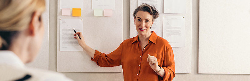 Female entrepreneur holding a pen and pointing to multiple sticky notes on the wall. Presenting the many ways having a business plan will benefit you as a business owner.