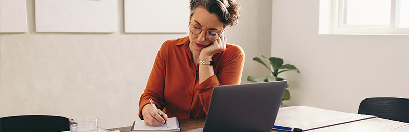 Female entrepreneur sitting at a desk in her home office. Jotting down notes on a notepad to fill in her internal business plan.