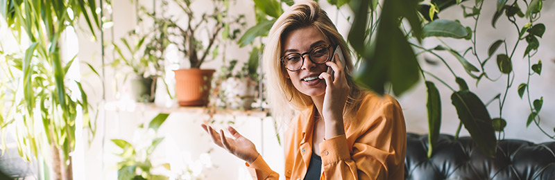 Female entrepreneur sitting outside surrounded by plants while talking on the phone asking a professional plan writer questions before hiring.