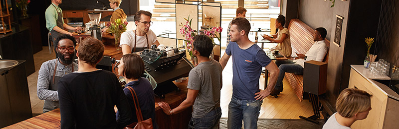 Group of seven individuals standing around inside of the entrance of a restaurant. Two are speaking with the owner, who just finished planning for his restaurant, preparing to order food.