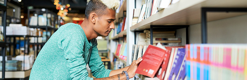 Male entrepreneur visiting a book store standing by a shelf reading through a book looking for business planning resources.